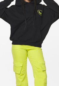 Zwarte hoodie met gele tekst en grafisch ontwerp, gecombineerd met felle gele cargobroek met meerdere zakken en een aansluitende taille.