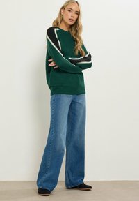 Maglione verde lavorato a maglia con dettagli a strisce bianche e nere, abbinato a jeans blu a gamba larga e scarpe nere.