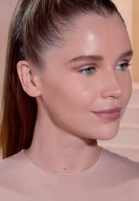 Close-up van een gezicht met een gladde huid, lichte make-up en een hoge paardenstaart. Het onderwerp draagt een doorzichtig, lichtroze top met een ronde hals.