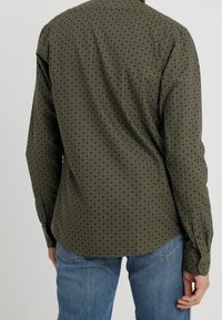 Oliwkowa koszula z długim rękawem w czarne kropki, z kołnierzykiem zapinanym na guziki i zaokrąglonym dołem, zestawiona z niebieskimi dżinsami. Materiał z domieszką bawełny.