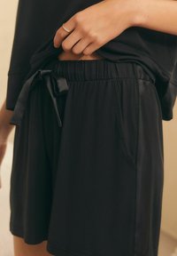 Un primo piano di una mano che tiene il bordo dei pantaloni corti neri, con un fiocco annodato, su uno sfondo soft.
