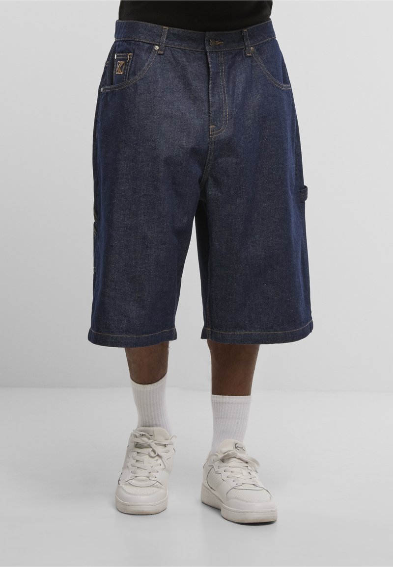 Bărbat purtând pantaloni scurți din denim închis la culoare, cu lungimea până la genunchi, șosete albe și adidași albi, stând în fața unui fundal simplu și luminos.