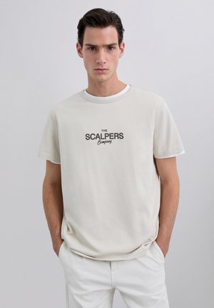 Camiseta de color crema con mangas cortas, que presenta un logo impreso en oscuro: "THE SCALPERS Company". Diseño sencillo de cuello redondo, textura de tela suave.