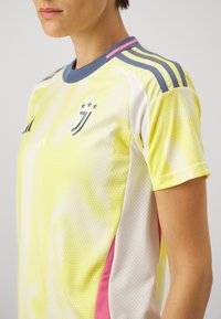 Personne portant un maillot de football Juventus jaune et blanc avec un col bleu foncé et des bandes sur les épaules, ainsi que des panneaux latéraux roses.