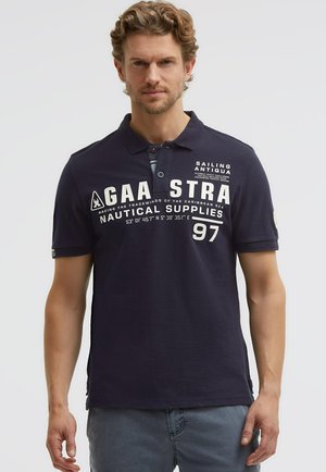 Mann mit hellbraunem Haar, der ein marineblaues Poloshirt mit weißem, nautisch inspiriertem Schriftzug und hellblaue Jeans trägt, steht vor einem einfarbigen Hintergrund.