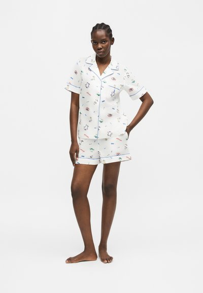 Γυναίκα στέκεται ξυπόλυτη φορώντας λευκό σετ πιτζάμες με κοντά μανίκια, πολύχρωμες μικρές στάμπες και μπλε λεπτομέρειες σε απλό φόντο.