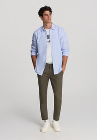 Camicia azzurro chiaro con bottoni sopra una maglietta bianca con grafica, abbinata a pantaloni slim-fit verdi oliva e sneakers bianche. Design semplice, look casual.