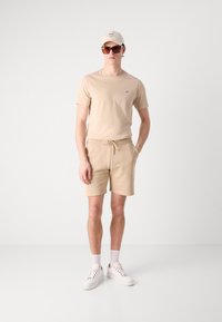 Beige kortærmet t-shirt og matchende shorts lavet af blødt stof. Hvide sneakers og en lys kappe med mørk tekst. Tilbehør inkluderer solbriller.