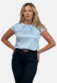 Jasnoniebieska satynowa bluzka z krótkim rękawem i plisowanym dekoltem, zestawiona z ciemnoniebieskimi dżinsami oraz czarnym paskiem z metalową klamrą logo.