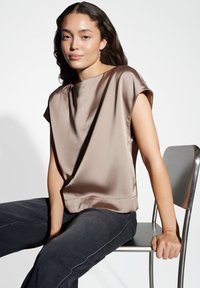 Top in satin marrone con maniche corte a cappuccio, vestibilità rilassata e collo alto. Modella seduta su una sedia di metallo, indossando jeans scuri.
