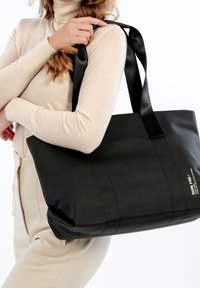 Kobieta w beżowym golfie i spodniach, trzymająca dużą czarną torbę na ramię z szerokimi paskami.