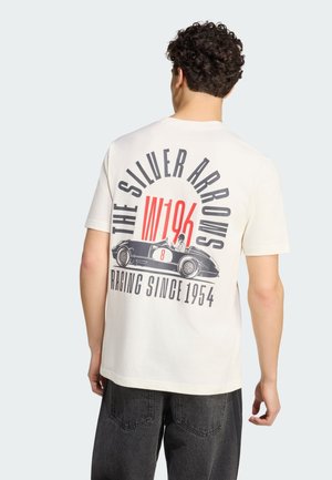 Mężczyzna nosi białą koszulkę z grafiką vintage samochodu wyścigowego, numerem 8 oraz napisem "The Silver Arrows" i "Racing since 1954" na plecach.