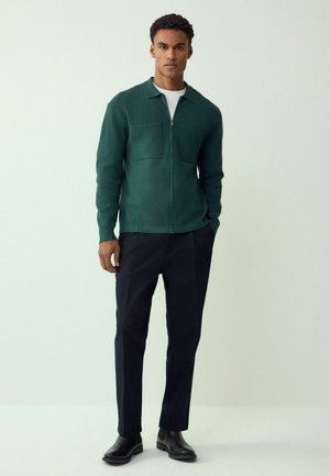 Green zip-up jacket with two front pockets, worn over a white shirt, paired with dark trousers and black boots. Simple design and soft texture.