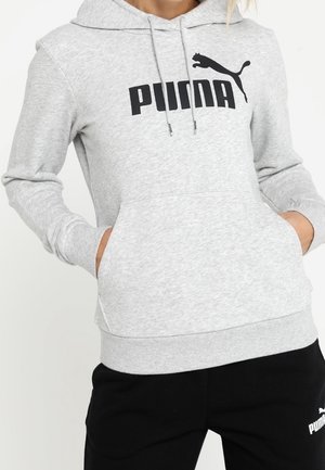 Osoba nosząca jasnoszarą bluzę z kapturem Puma z czarnym logo, z rękami w przedniej kieszeni, w zestawie z czarnymi spodniami dresowymi Puma.