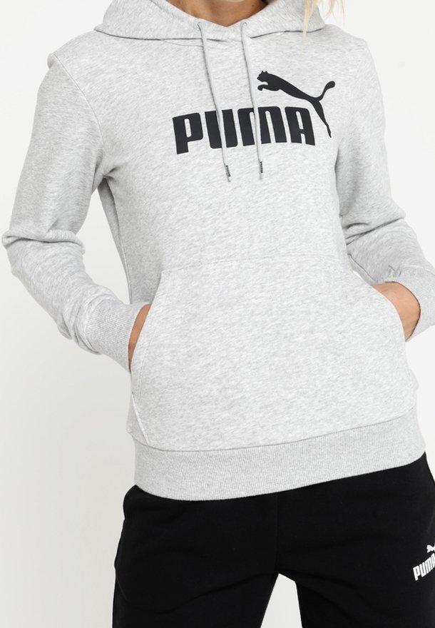 Personne portant un sweat à capuche Puma gris clair avec logo noir, mains dans la poche avant, associé à un pantalon de survêtement noir Puma.