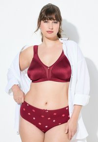 Bordeaux kant beha met brede bandjes en laag uitgesneden slip met bloemmotief, beide met een gladde textuur. Het model draagt een open witte shirt.
