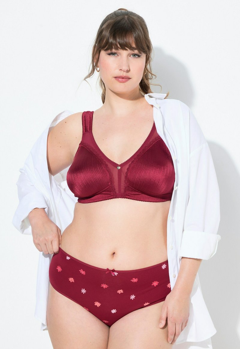 Bordeaux kant beha met brede bandjes en laag uitgesneden slip met bloemmotief, beide met een gladde textuur. Het model draagt een open witte shirt.
