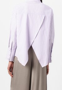 Vue de dos d'une personne portant une chemise blanche à fines rayures verticales avec un dos ouvert superposé, associée à un pantalon beige ample.