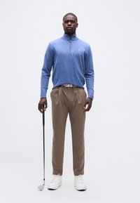 Άνδρας με μπλε πουλόβερ με φερμουάρ μέχρι τη μέση, μπεζ παντελόνι και λευκά αθλητικά παπούτσια κρατώντας ένα μπαστούνι γκολφ, στέκεται μπροστά σε ένα απλό φόντο.