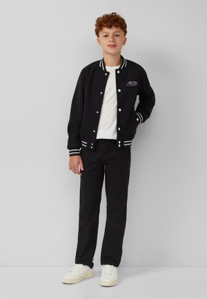 Un garçon aux cheveux bouclés et roux se tient contre un mur gris, portant une veste varsity noire, un T-shirt blanc, un pantalon noir et des baskets blanches.