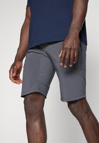 Graue Sportshorts mit glatter Textur, einer Reißverschlusstasche und eckigem Design. Tragen mit einem dunkelblauen Shirt, das einen taillierten Schnitt zeigt.