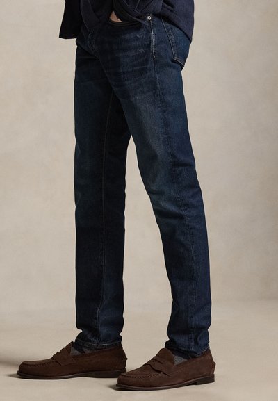 Vyras dėvi tamsiai mėlynas džinsines kelnes ir rudus zomšinius batelius, stovintis neutraliame fone.