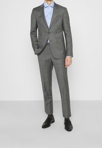 Szary garnitur szyty na miarę z dwoma guzikami, klapami z wycięciem oraz spodniami z prostym przodem, zestawiony ze światło niebieską koszulą i czarnymi mokasynami.