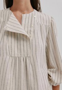 Beige linnen blouse met verticale marineblauwe strepen, voorzien van een sjaalkraag, drie knopen, geplooide details en soepele mouwen.