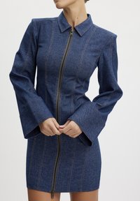 Jeansklänning med en figurnära silhuett, hög krage, långa utsvängda ärmar och dragkedja fram. Har kontrasterande sömmar nedanför framsidan.