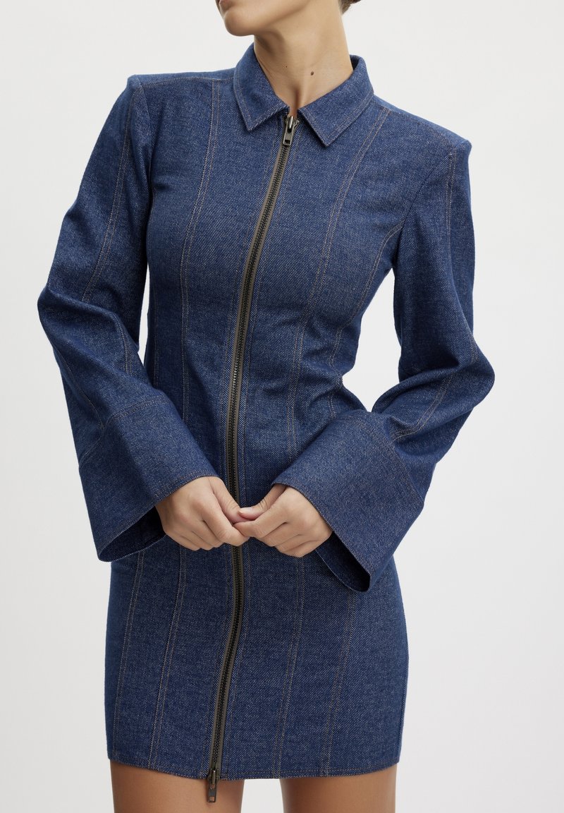 Jeansklänning med en figurnära silhuett, hög krage, långa utsvängda ärmar och dragkedja fram. Har kontrasterande sömmar nedanför framsidan.