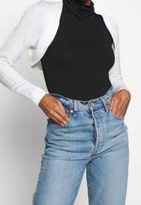 Schwarzer Rollkragenbodysuit unter einer weißen kurzen Strickjacke, kombiniert mit hochgeschnittenen, hellblauen Jeans mit ausgewaschenem Look und Fronttaschen.