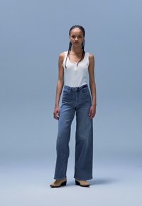 Lys blå denimjeans med høy midje og løs passform. Kombinert med en hvit ermeløs topp med knappedetalj. Sorte høyhælte sko.