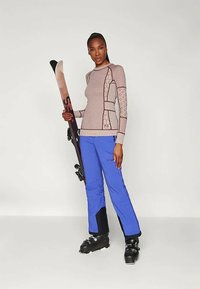 Αθλητική ενδυμασία σκι που περιλαμβάνει μια μπεζ μακρυμάνικη μπλούζα με σχέδια και ραφές ριμπ, καθώς και μπλε παντελόνια σκι. Τα αξεσουάρ περιλαμβάνουν μαύρες μπότες σκι και σκι.