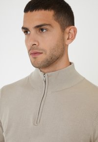 Beige knit sweater with a high collar and half-zip closure. Features ribbed texture around the collar and cuffs.
