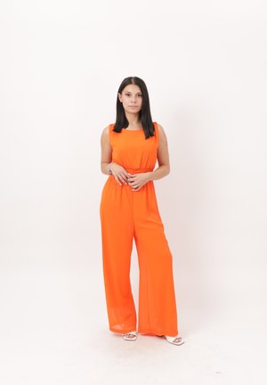 Ung kvinde med glat mørkt hår iført en knaldrød orange ærmeløs overall og hvide åbne tå-hæle, der står mod en ensfarvet lys baggrund.