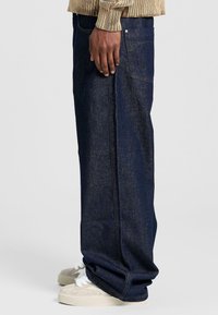Jeans in denim blu con taglio dritto, tasche laterali e dettagli di cucitura discreti, indossati su sneakers chiare.