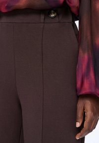 Bruine stoffen broek met een gladde textuur, voorzien van een enkele knoopsluiting en zijzakken, gecombineerd met een meerkleurige blouse.