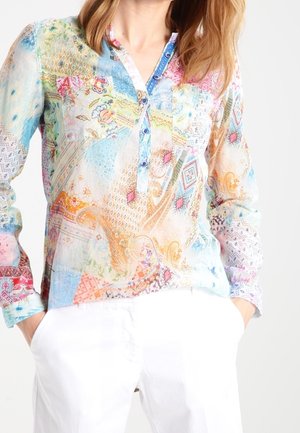 Femme portant un chemisier à manches longues coloré et à motifs avec un col boutonné, assorti d'un pantalon blanc, les mains dans les poches.