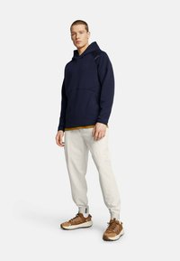 Marineblauer Kapuzenpullover mit einem kontrastierenden senfgelben Saum, kombiniert mit cremfarbigen Jogginghosen und braunen Sneakern mit weißen Akzenten. Insgesamt glatte Textur.