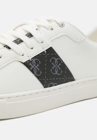 Fehér bőr sneaker fekete texturált oldalpanelekkel, amelyek finom szürke logópattintás mintázattal rendelkeznek. Fehér fűző és strapabíró gumi talp.