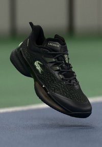Svart atletisk sko med et teksturert stoffoverdel, grønne detaljer og et karakteristisk logo. Har en polstret tunge og fleksibel såle.