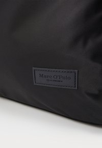 Czarna torba z nylonu o teksturowej powierzchni, z prostokątną skórzaną metką z wytłoczonym napisem "Marc O'Polo" oraz "EST. W Sztokholmie."