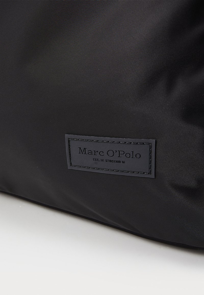 Czarna torba z nylonu o teksturowej powierzchni, z prostokątną skórzaną metką z wytłoczonym napisem "Marc O'Polo" oraz "EST. W Sztokholmie."