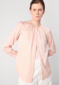 Blusa color pescoce con maniche lunghe e chiusura a bottone, caratterizzata da arricciature all'escursione e tessuto leggero e morbido.