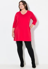 Rode tuniekjurk met een V-hals en driekwartmouwen, gecombineerd met zwarte legging en enkellaarsjes. Gladde textuur; minimaal ontwerp.
