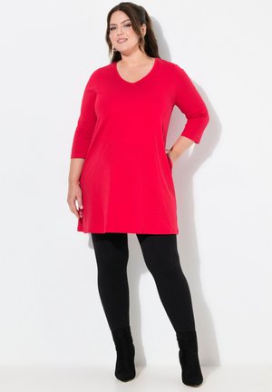 Rotes Tunika-Kleid mit V-Ausschnitt und Dreiviertelärmeln, kombiniert mit schwarzen Leggings und Stiefeletten. Glatte Textur; minimales Design.
