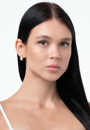 Orecchini in oro con una pietra bianca a forma di pera, texture liscia e design minimalista. Contrasto con capelli scuri e sfondo neutro.