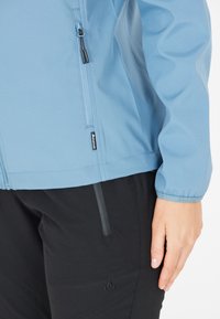 Veste soft-shell bleu clair avec une poche zippée et des poignets élastiques, associée à un pantalon noir et un logo visible sur la jambe.
