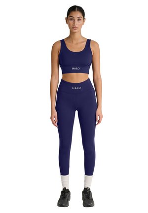 Kvinde iført matchende mørkeblå crop top og leggings med "HALO" logo, hvide sokker og sorte træningssko, stående oprejst.