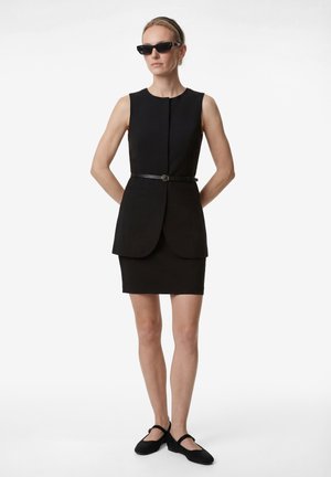 Mujer que lleva un vestido negro sin mangas con cinturón, bailarinas negras Mary Jane, gafas de sol oscuras y el pelo recogido, de pie con las manos detrás de la espalda.